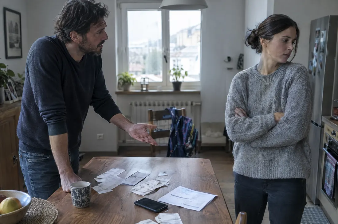 Un uomo e una donna discutono in cucina, ai due lati di un tavolo con scontrini e fatture, mentre lo zainetto di un figlio è appeso a una sedia (immagine illustrativa)