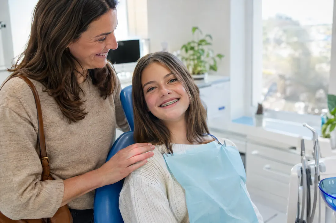 Una ragazza di dodici anni con l'apparecchio ai denti sorride sulla poltrona dell'ortodontista, accanto alla madre